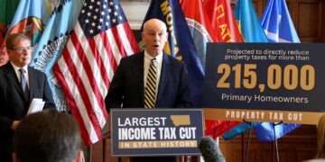 Speaker at podium flanked by multiple flags during a press conference or rally about tax cuts and homeowners, with a large banner in the foreground reading 'Largest income tax cut'.