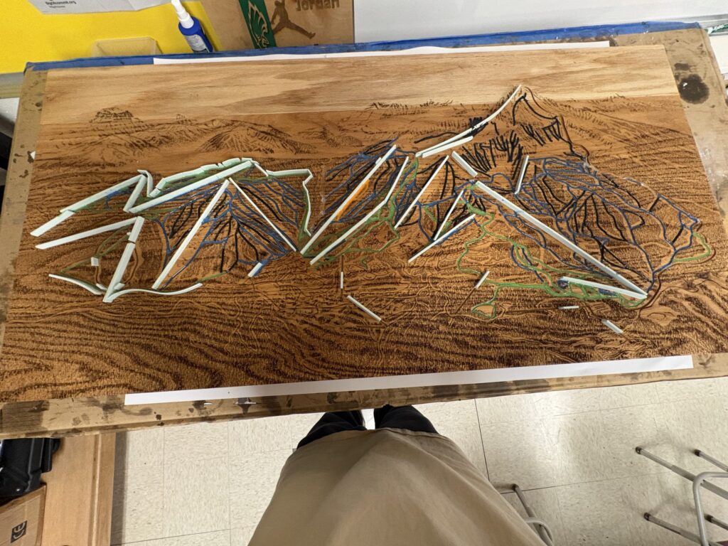 Art project on a wooden table featuring tangled blue and green strings with scattered white plastic rods and fragments nearby.