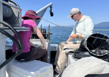 Two people sit on a small boat by the water, fishing with rods and gear under a clear blue sky.