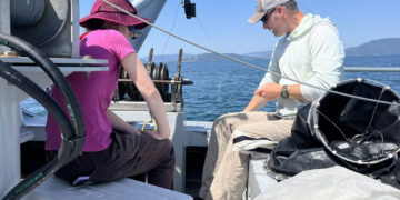 Two people sit on a small boat by the water, fishing with rods and gear under a clear blue sky.