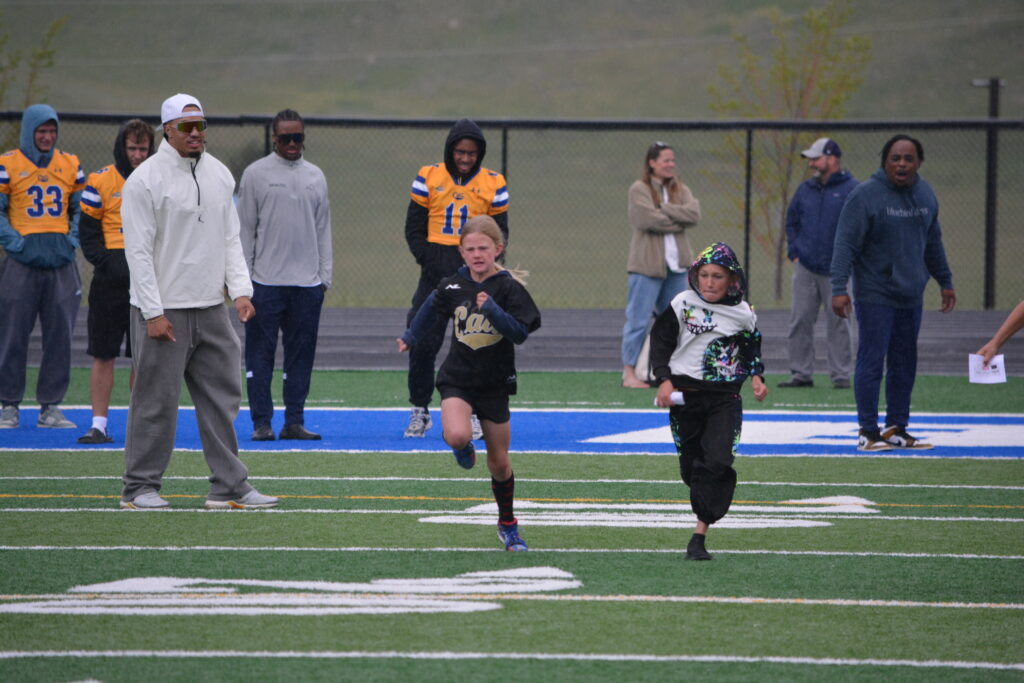Children sprinting on a football field as adults and coaches watch from the sidelines behind a chain-link fence on a cloudy day.