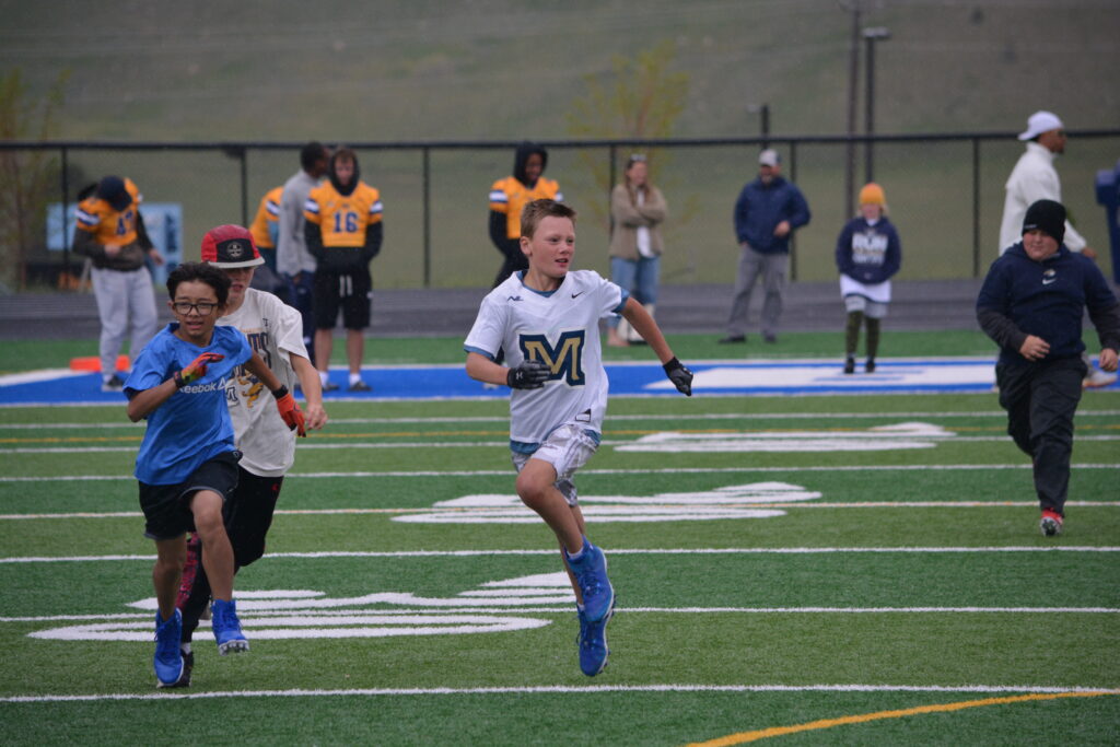 Young boys sprint on a football field during a race, with players and spectators in the background on a rainy day.