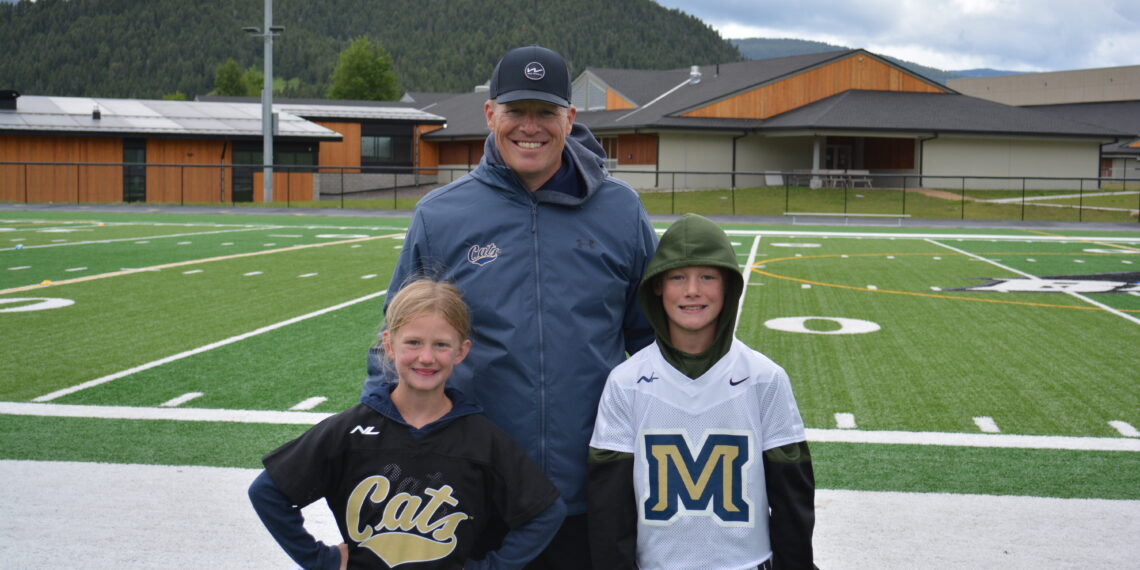 Man in a blue jacket with two children posing on a football field, with hills and school buildings in the background.