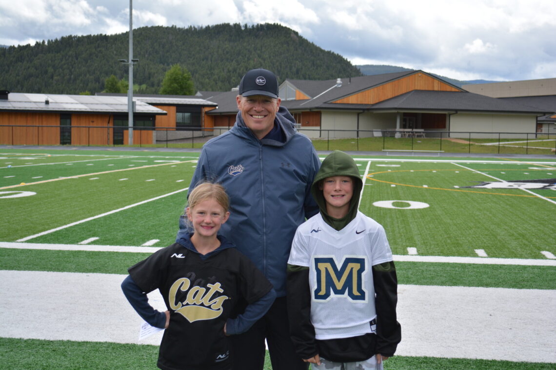 Man in a blue jacket with two children posing on a football field, with hills and school buildings in the background.