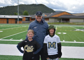 Man in a blue jacket with two children posing on a football field, with hills and school buildings in the background.