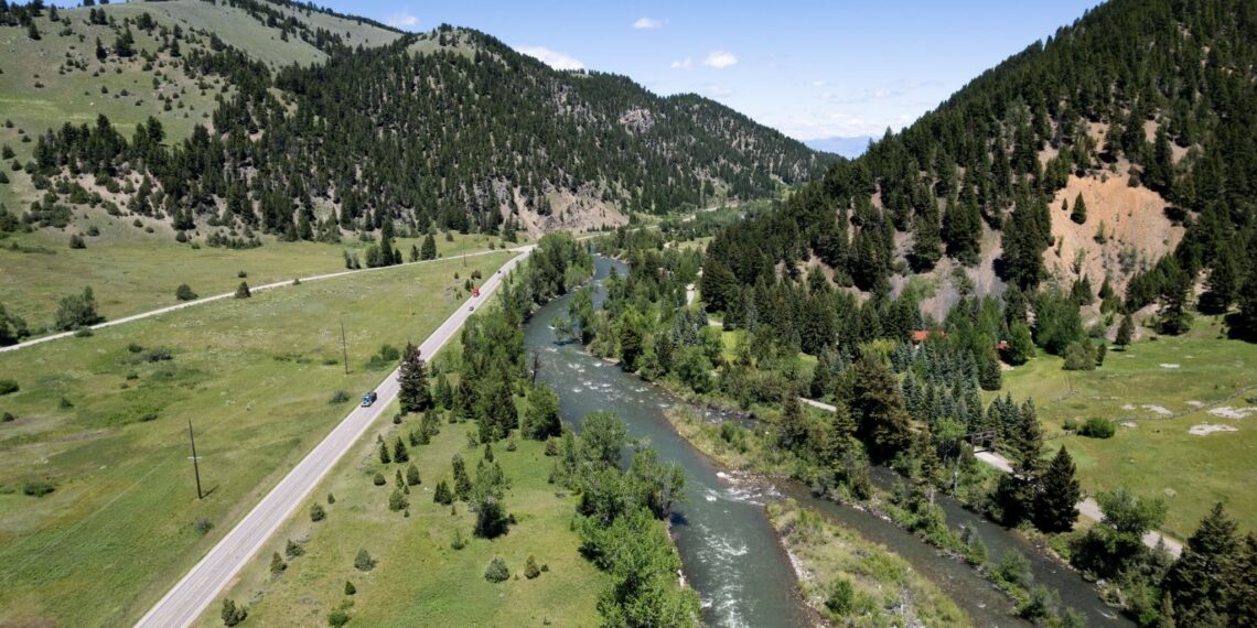 Panoramic valley with a winding river flanked by forested hills and a straight road running parallel to the water under a clear blue sky.