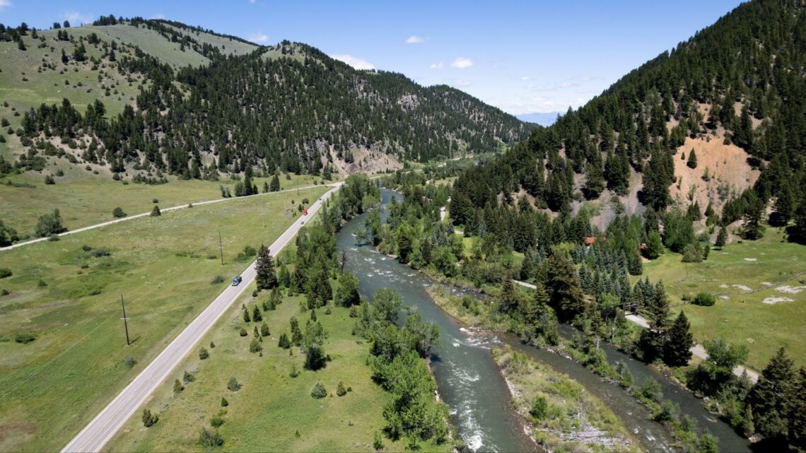 Panoramic valley with a winding river flanked by forested hills and a straight road running parallel to the water under a clear blue sky.