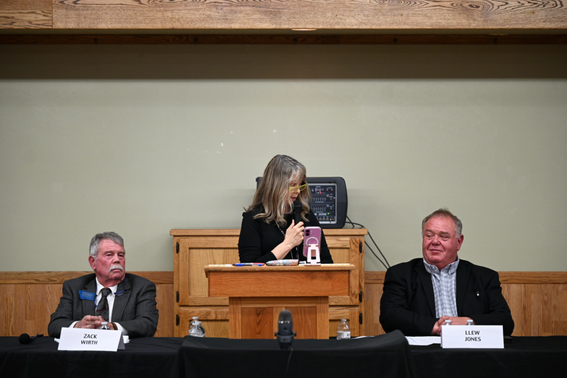 Panel of three people seated at a wooden desk; a woman in the center speaks into a mic while holding a pink phone, flanked by two men in suits.