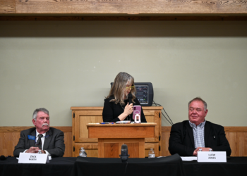 Panel of three people seated at a wooden desk; a woman in the center speaks into a mic while holding a pink phone, flanked by two men in suits.