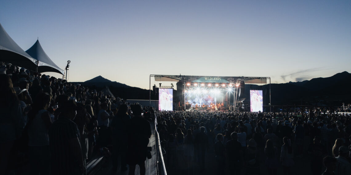 Large crowd at an outdoor music festival at dusk, with a brightly lit stage and tents on the left.