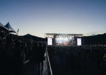 Large crowd at an outdoor music festival at dusk, with a brightly lit stage and tents on the left.