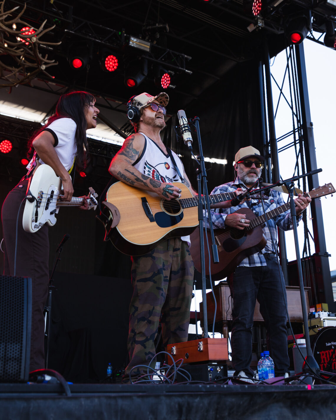 Three musicians perform on an outdoor stage; center man sings into a microphone with an acoustic guitar, wearing a camo cap and sunglasses.