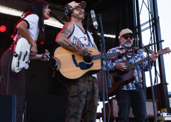 Three musicians perform on an outdoor stage; center man sings into a microphone with an acoustic guitar, wearing a camo cap and sunglasses.