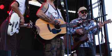 Three musicians perform on an outdoor stage; center man sings into a microphone with an acoustic guitar, wearing a camo cap and sunglasses.
