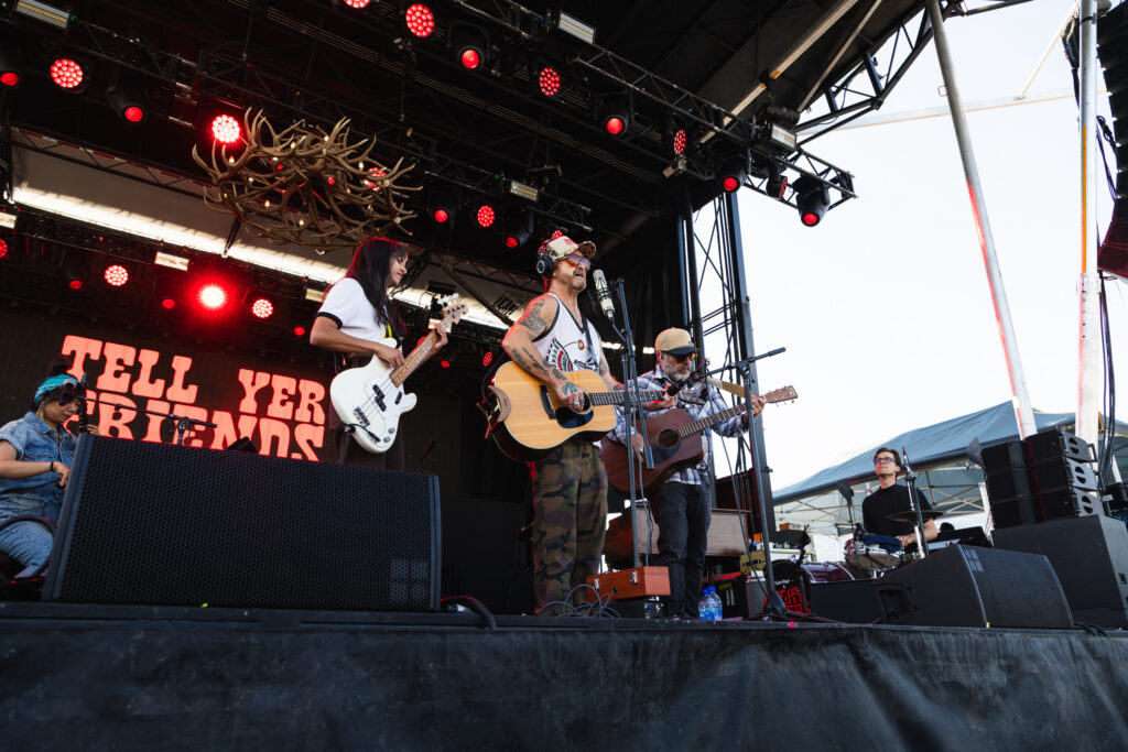 Band performing on an outdoor stage with guitars, drummer in back, and a decorative antler chandelier overhead while red stage lights shine down.