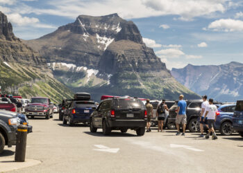 Crowded mountain parking lot with SUVs and trucks, hikers walking between vehicles, and rocky peaks in the background.