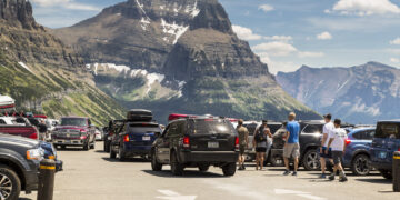 Crowded mountain parking lot with SUVs and trucks, hikers walking between vehicles, and rocky peaks in the background.