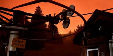Silhouetted telecom equipment and satellite dishes against an orange smoky sky, with a small building and a 'Keep Tips Up' sign nearby.