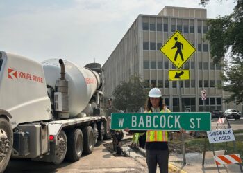 Construction worker in hard hat holding a street sign on a closed sidewalk near a concrete mixer truck and a yellow pedestrian crossing sign.