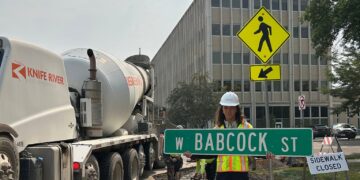 Construction worker in hard hat holding a street sign on a closed sidewalk near a concrete mixer truck and a yellow pedestrian crossing sign.