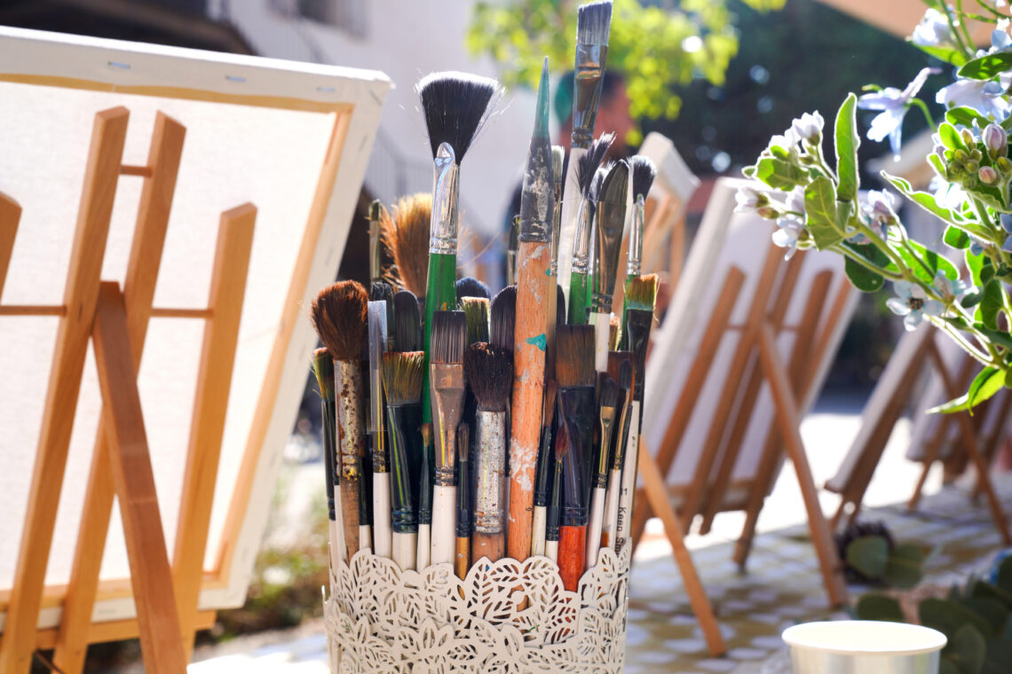 Cluster of paintbrushes in a white decorative holder outdoors, with easels in the background and white flowers nearby.