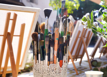 Cluster of paintbrushes in a white decorative holder outdoors, with easels in the background and white flowers nearby.