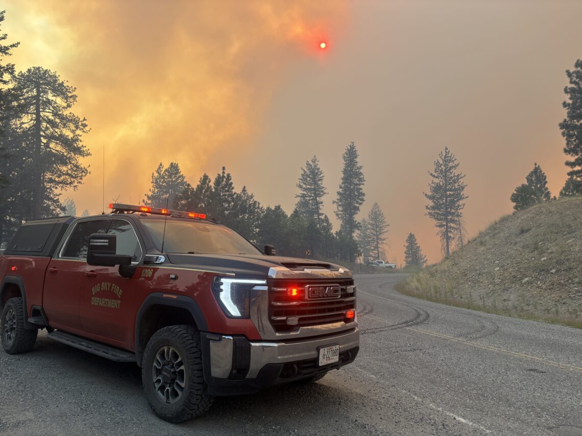 Fire department pickup with emergency lights on a smoky road, forest fire in the background.