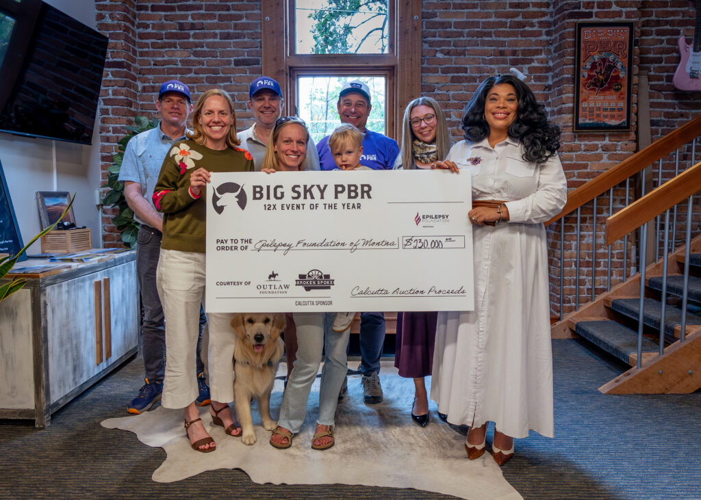 Group of adults posing indoors behind a large ceremonial check, with a golden retriever sitting in front, in a brick-walled space. The check reads 'BIG SKY PBR 12x Event of the Year'.
