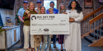 Group of adults posing indoors behind a large ceremonial check, with a golden retriever sitting in front, in a brick-walled space. The check reads 'BIG SKY PBR 12x Event of the Year'.