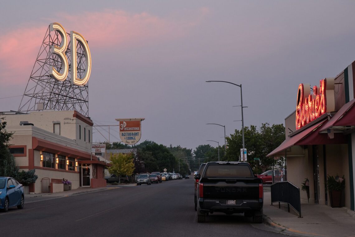 Evening street scene with a large neon '3D' sign atop a framework on the left, and a red neon 'Diners' sign on the right, with parked cars along the street.