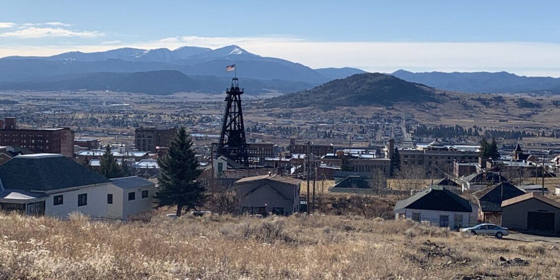 Mining headframe with an American flag rising over a town, with snow-dusted mountains in the distance under a clear blue sky(panoramic view).