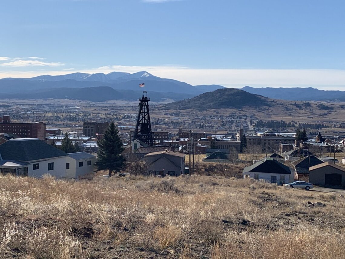 Mining headframe with an American flag rising over a town, with snow-dusted mountains in the distance under a clear blue sky(panoramic view).