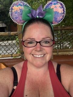 Smiling woman wearing a purple-green sparkly mouse ears headband, red-framed glasses, and a red tank top, outdoors near a wooden fence and greenery.