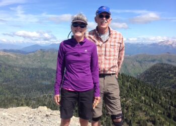 Two hikers standing on a rocky mountain summit with a forested valley and blue sky behind them.