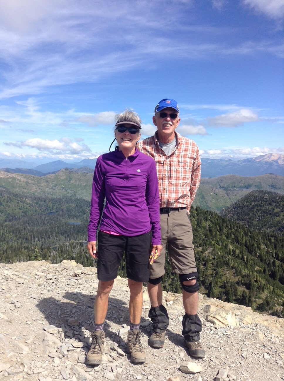 Two hikers standing on a rocky mountain summit with a forested valley and blue sky behind them.
