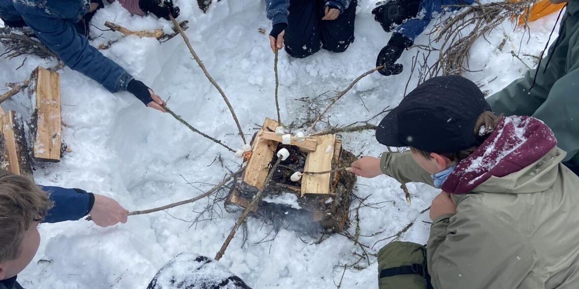 Group of children in winter coats and hats gathered around a wooden fire pit in the snow, arranging sticks and logs.