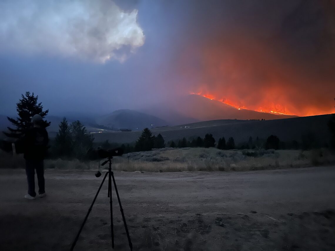 Silhouette of a person with a camera on a tripod watching a hillside wildfire with bright orange flames and smoke at dusk.