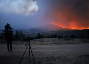 Silhouette of a person with a camera on a tripod watching a hillside wildfire with bright orange flames and smoke at dusk.