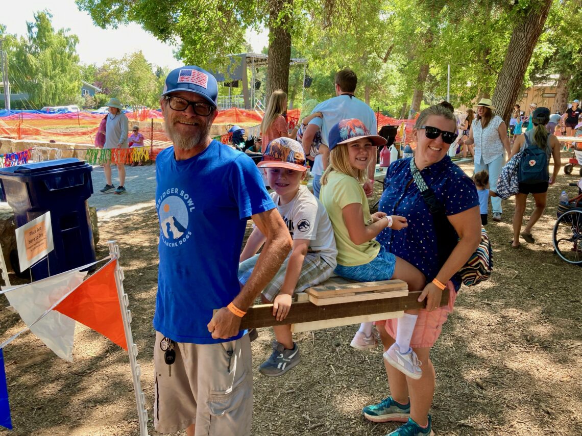 Group of people at an outdoor festival around a wooden obstacle; a man in a blue shirt and cap smiles near kids by a wooden beam.