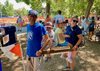 Group of people at an outdoor festival around a wooden obstacle; a man in a blue shirt and cap smiles near kids by a wooden beam.
