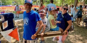 Group of people at an outdoor festival around a wooden obstacle; a man in a blue shirt and cap smiles near kids by a wooden beam.