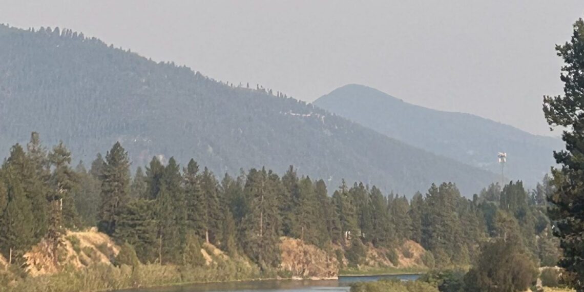 River flows through a forested valley with distant blue mountains under a hazy sky.
