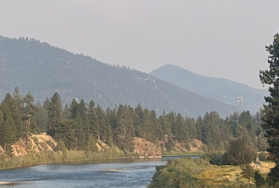 River flows through a forested valley with distant blue mountains under a hazy sky.