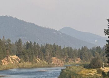 River flows through a forested valley with distant blue mountains under a hazy sky.