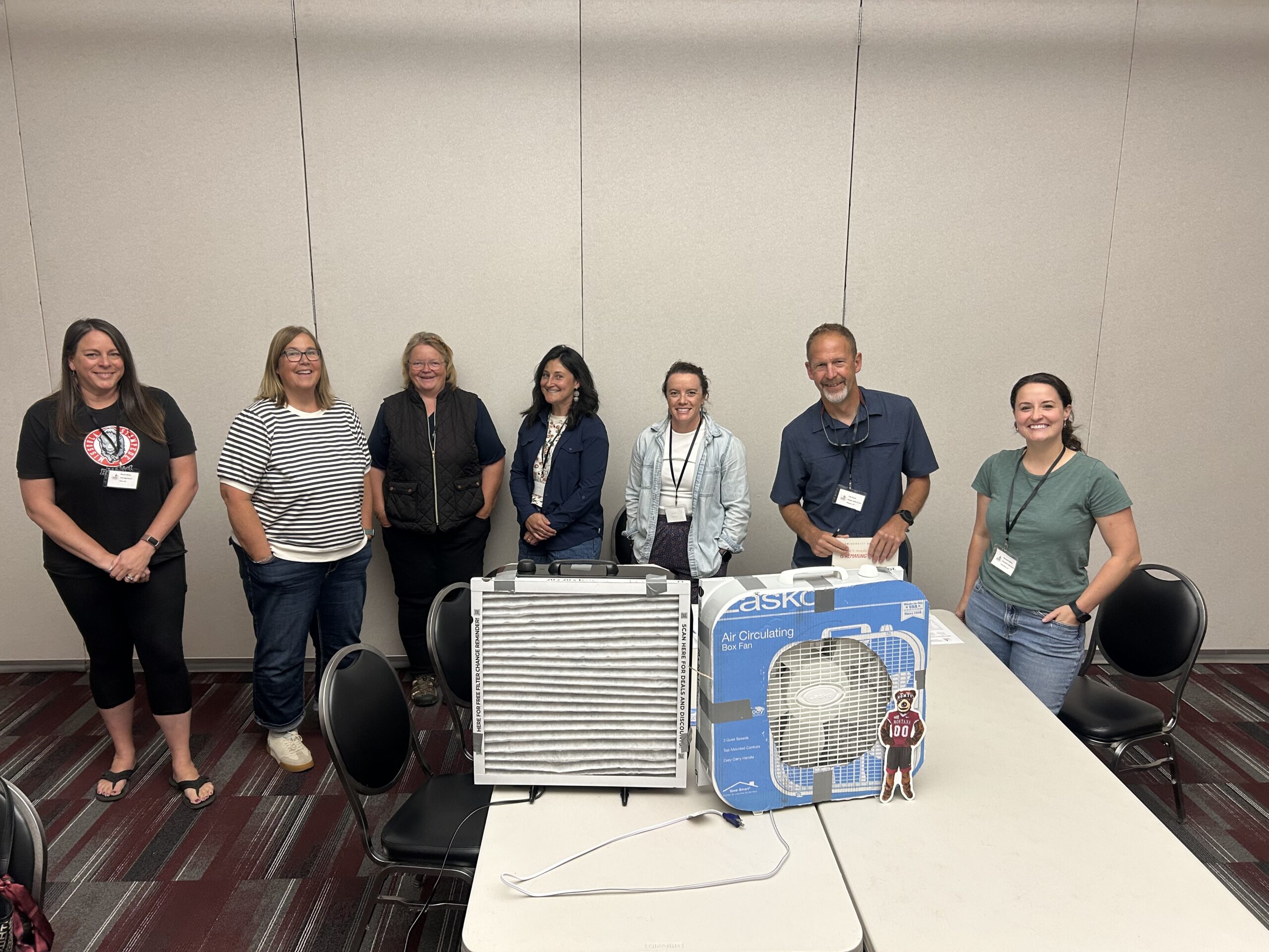 Seven adults stand in a line indoors behind a table with two large boxed air circulators, posing for a group photo in a conference room.