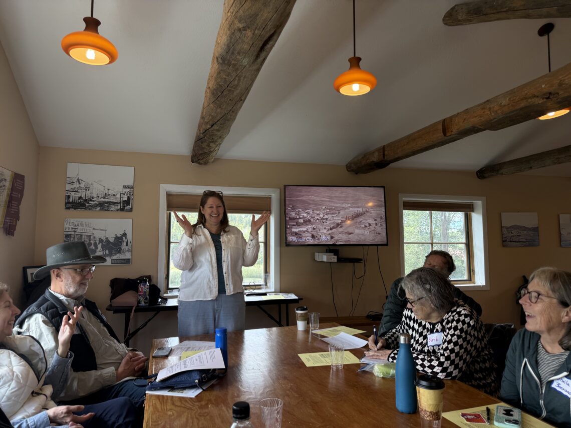 Woman standing at head of a wooden conference table presenting to a seated group in a room with exposed wooden beams and warm lighting.