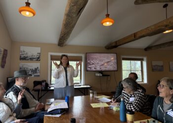 Woman standing at head of a wooden conference table presenting to a seated group in a room with exposed wooden beams and warm lighting.