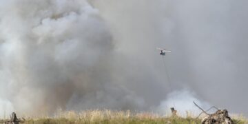 Helicopter with a long line and water bucket hovering over a smoky field during a wildfire.