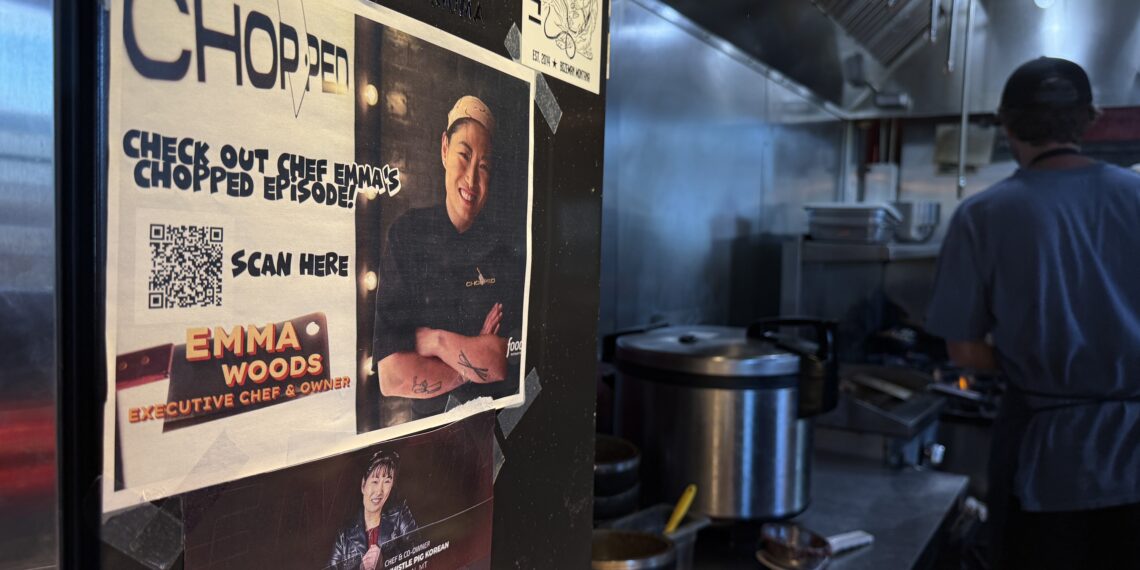 Promotional posters and stickers plastered on a black wall in a busy kitchen, with a person in a blue shirt working near stainless steel appliances in the background.