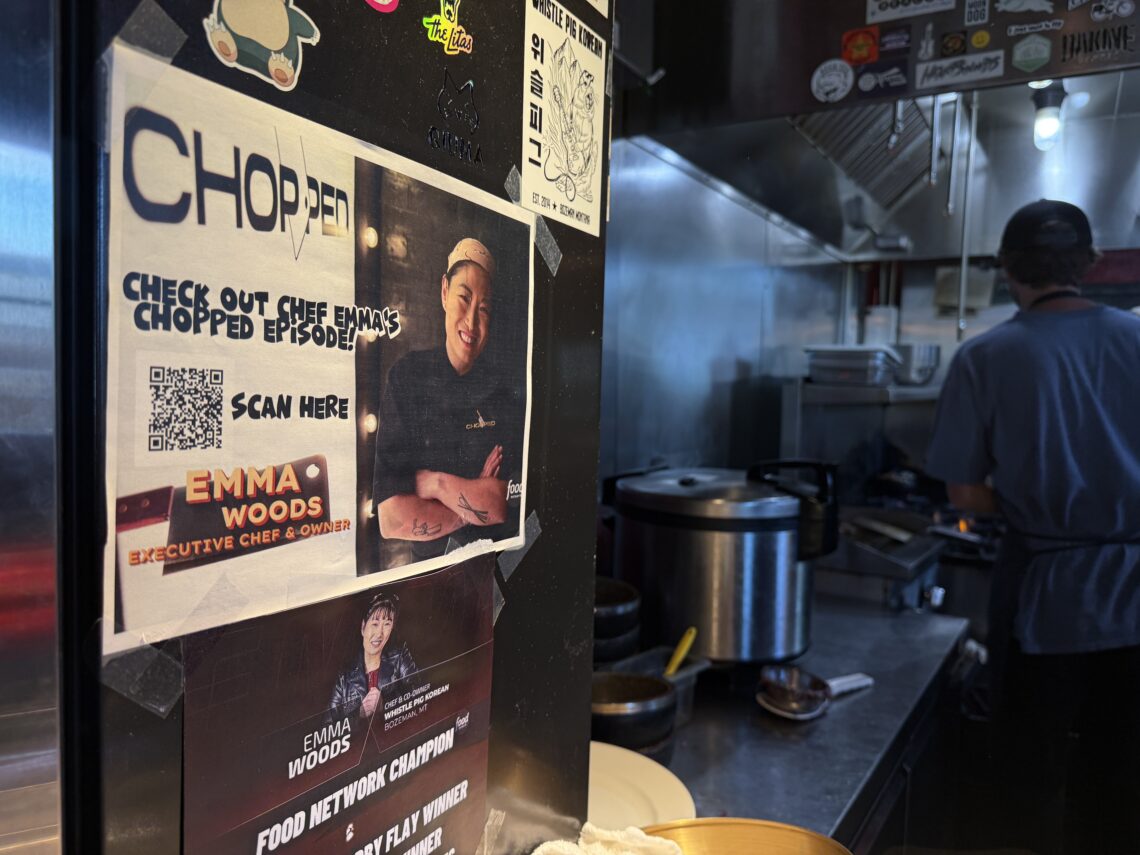 Promotional posters and stickers plastered on a black wall in a busy kitchen, with a person in a blue shirt working near stainless steel appliances in the background.